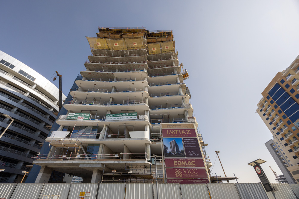 Cove Edition I By Imtiaz 1 A multi-story building under construction with scaffolding and safety signs, surrounded by modern buildings.